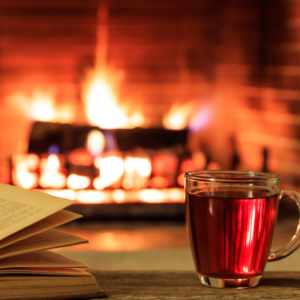 a cup of hot cider next to an open book with a lighted fireplace in the background